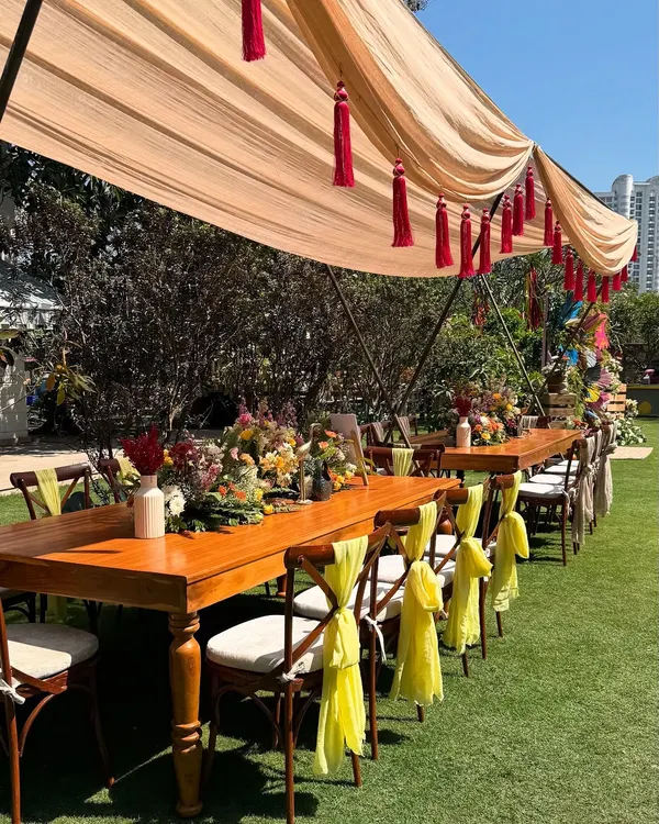 A beautiful long table set for an outdoor Holi lunch. The wooden table is decorated with a runner of fresh, colorful flowers, and the chairs are adorned with bright yellow fabric ties, adding to the festive atmosphere.