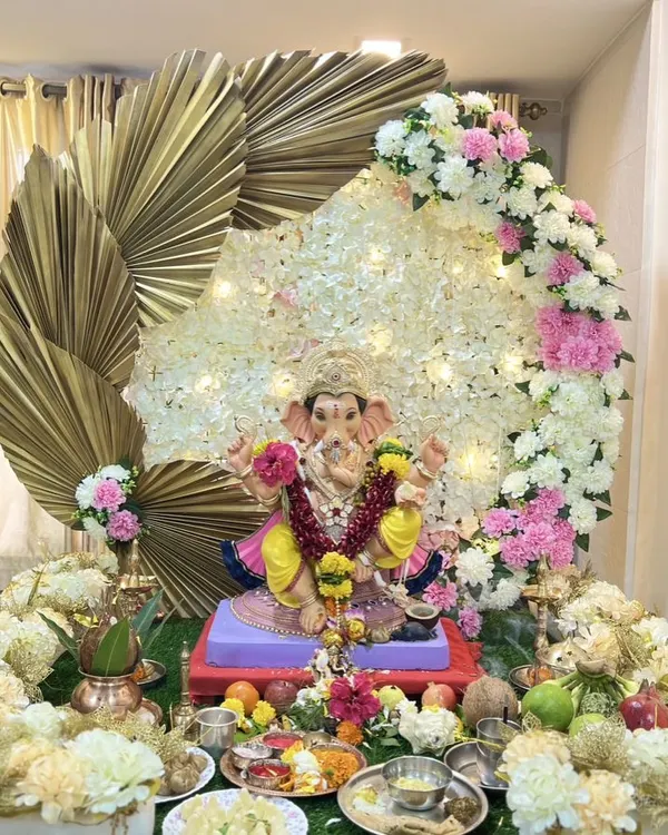 A stunning Ganpati decoration featuring a backdrop of white flowers and fairy lights, framed by large golden palm leaves for a modern, divine look.