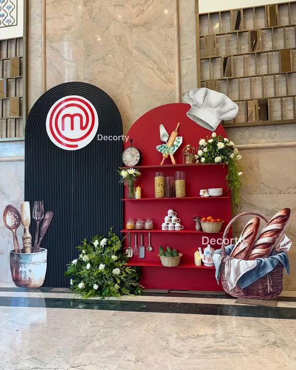 I create bold, thematic photo opportunities like this one for my Masterchef events. The setup includes a custom red shelving unit with cooking ingredients, a branded backdrop, and fun props like a chef's hat.