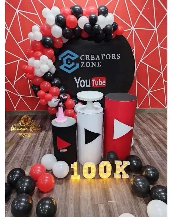 This is the full setup for the Creators Zone 100k subscriber celebration. It includes a branded backdrop, YouTube-themed pedestals, and marquee lights, all framed by a red, black, and white balloon garland.