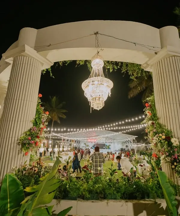 A grand entrance arch designed with stately white pillars, adorned with floral arrangements and magnificent hanging chandeliers. This entrance opens up to an outdoor reception, promising a night of elegance and celebration.