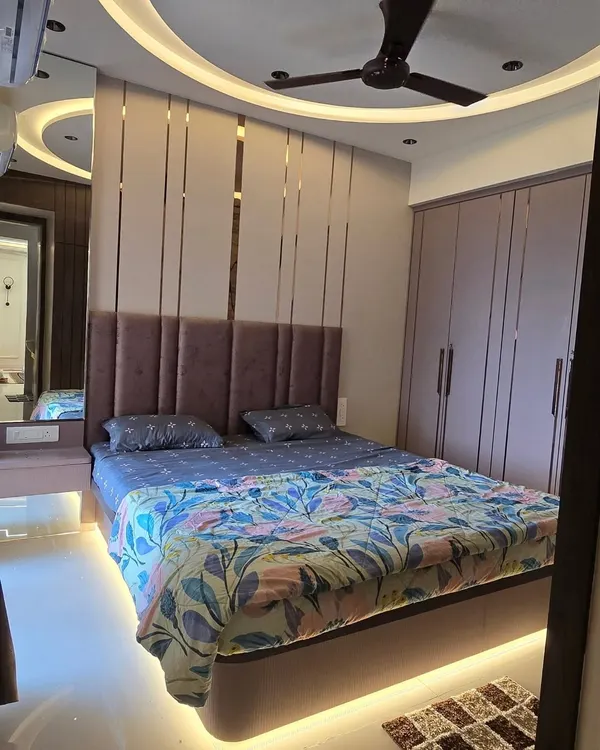 This master bedroom combines layers of texture for a cozy feel. The design features a plush upholstered headboard, elegant gold-tone metal inlays, and a curved false ceiling with concealed lighting.