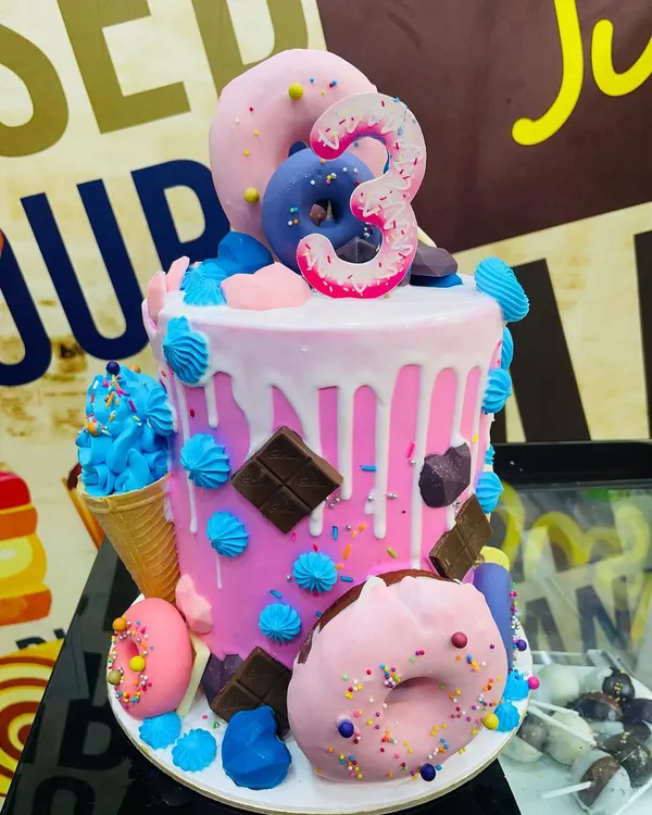 A vibrant, multi-layered drip cake perfect for a child's third birthday celebration. This custom creation features bright pink buttercream, a white chocolate drip, and is generously decorated with an assortment of treats including sprinkle-covered donuts, chocolate bars, and a miniature ice cream cone.