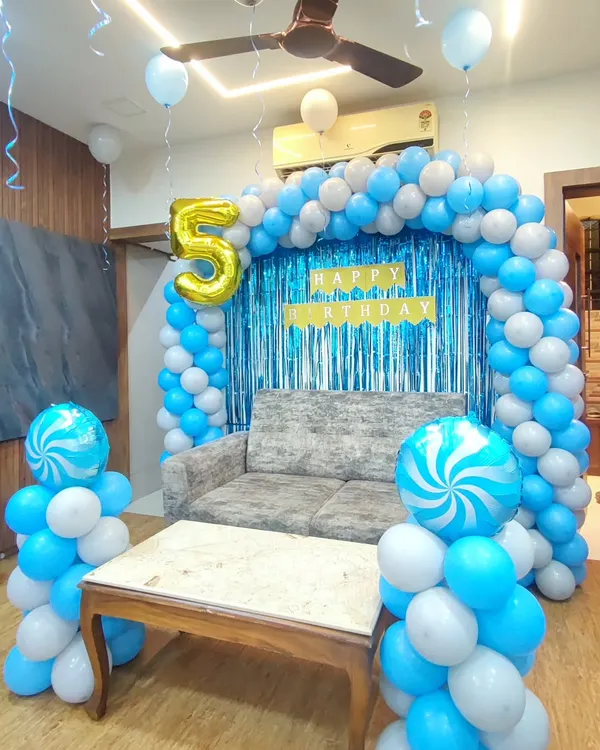 A simple and elegant birthday setup for a fifth birthday using a blue and white color scheme. The decoration features a balloon arch framing a blue shimmer backdrop, with two candy-swirl foil balloons on columns.