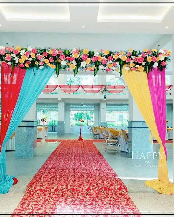 A bright and cheerful entranceway for a wedding hall, decorated with colorful drapes in red, blue, yellow, and pink. A lush floral arrangement at the top completes this festive welcome for your guests.