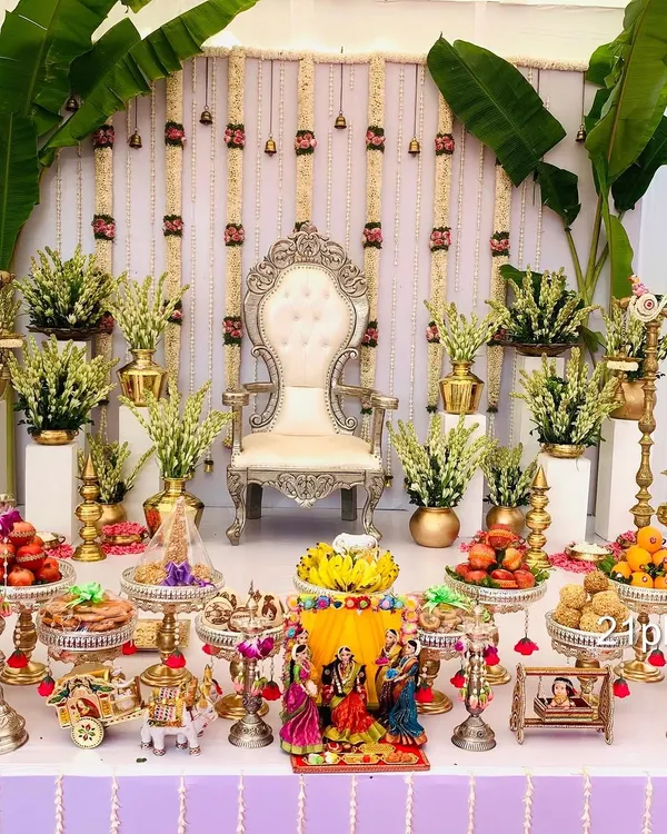 An elegant and classic Seemantham setup featuring a silver throne, fresh jasmine and tuberose flower arrangements, and large banana plants for an authentic traditional feel. The foreground is filled with auspicious items and doll displays.