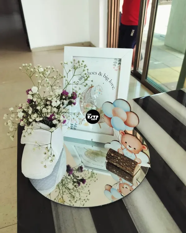 This sweet centerpiece captures the essence of a teddy bear theme. It features a cute bear and balloon cutout, a framed print with a heartfelt quote, and a baby shoe holding delicate flowers, all arranged on a mirrored tray.