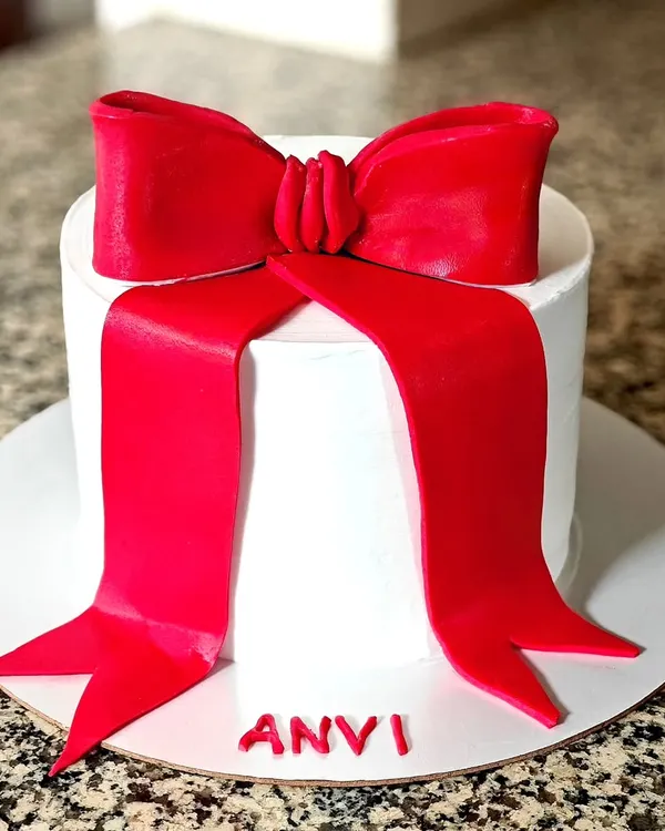 The core of elegance is simplicity. This cake features a clean white buttercream finish and a striking, handcrafted red fondant bow, personalized with the name 'Anvi'.