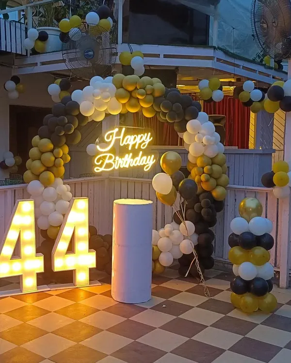 A stylish outdoor terrace decoration for a 44th birthday. I used a black, yellow, and white balloon arch, a 'Happy Birthday' neon sign, and large marquee numbers to create a festive photo corner.