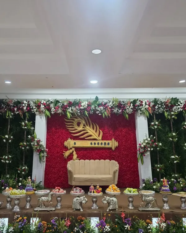 A unique stage design with a Krishna theme, featuring a large golden flute and peacock feather against a wall of red roses.