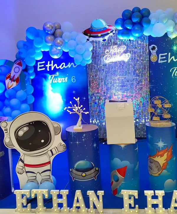 A futuristic space theme with cool blue lighting and a shimmering sequin wall. The setup is filled with astronaut and rocket cutouts for a cosmic feel.