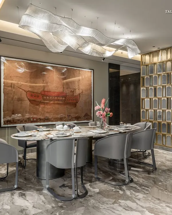 This dining area in a Shivaji Park residence is envisioned as a central anchor of the home. A contemporary theme with a nod to tradition is set by a statement wall etched with ancient Indian texts, while a dramatic wavy light fixture floats above the marble table.