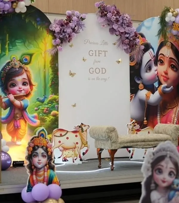 A divine Krishna-themed baby shower setup in shades of purple and gold. The backdrop features an illustration of baby Krishna, with a sign that reads "Precious Little Gift from God is on the way," and is framed by a beautiful balloon arch.