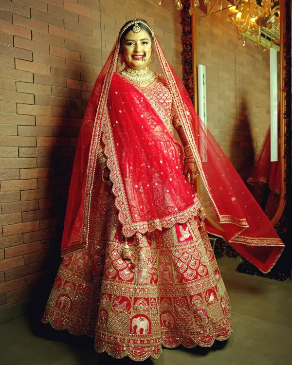 A classic full-length bridal portrait against a brick wall, capturing the bride's joyful smile and the intricate details of her red wedding lehenga.