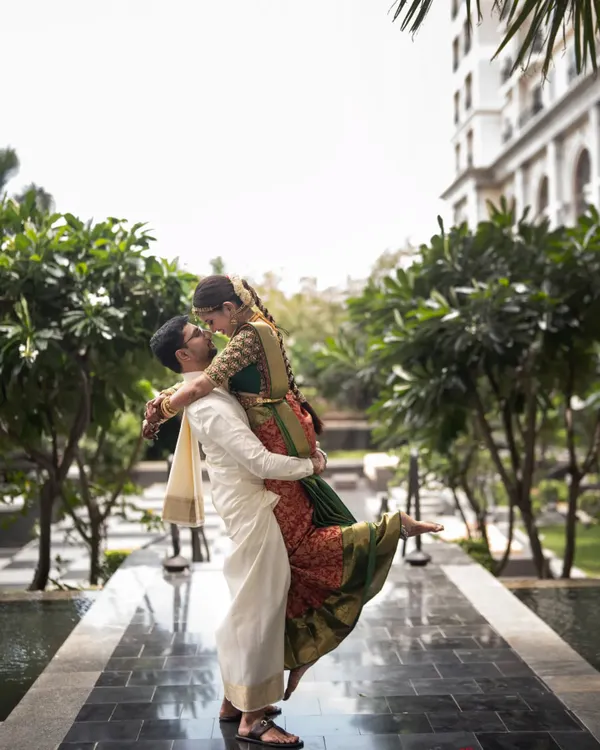 A playful and romantic shot of the couple. The groom lifts his bride, showing the beautiful drape of her traditional red and green Kanjivaram saree against his classic white veshti. It’s a moment of pure happiness.