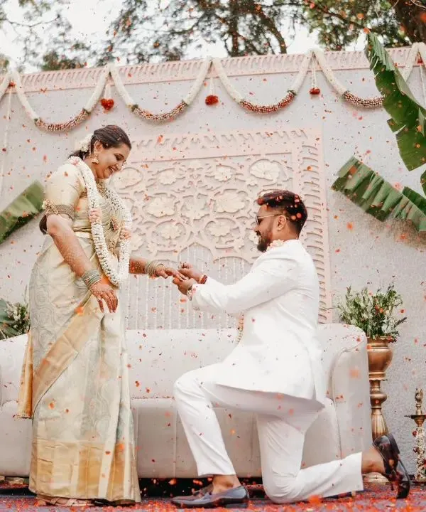 A beautiful proposal moment during a traditional engagement ceremony. The backdrop is a pristine white panel with intricate carvings, flanked by banana plants and traditional brass lamps, creating a purely elegant and culturally rich setting.