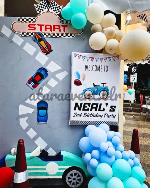 The race begins here! A "Welcome to Neal's 2nd Birthday Party" sign sets the stage, complete with a custom racetrack backdrop, car cutouts, and a blue balloon garland.