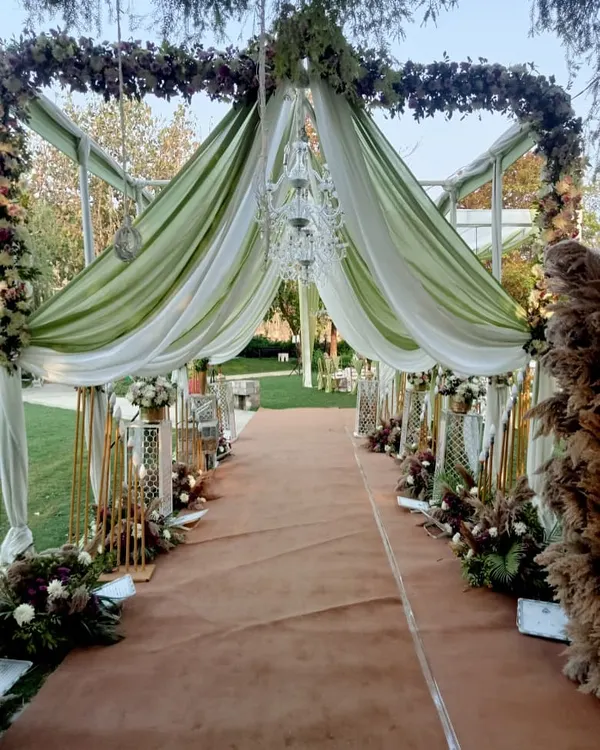 A breathtaking entrance for a day wedding, with a walkway covered in green and white drapes, floral arrangements, and elegant chandeliers.