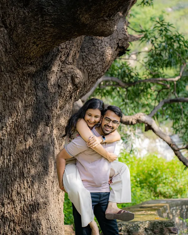 A playful and happy moment with a piggyback ride under a large, beautiful tree. This candid shot from an outdoor pre-wedding session is full of natural joy and laughter.