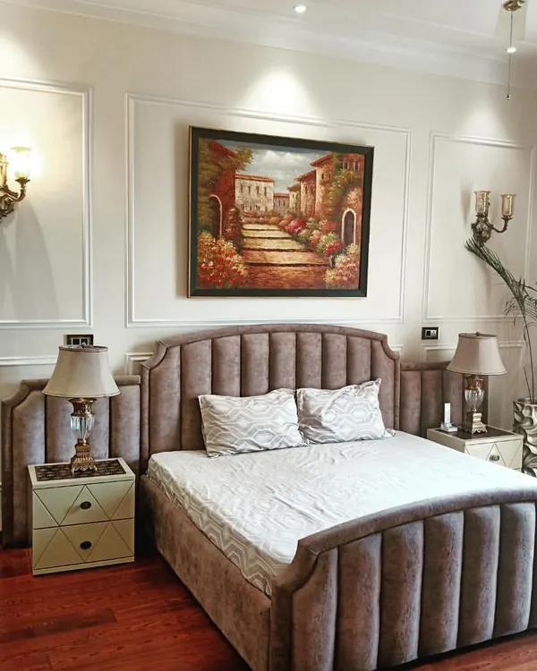 A master bedroom designed with classic elegance. It features a channel-tufted upholstered bed, traditional wall moulding, classic sconces, and rich wooden flooring for a timeless and cozy feel.
