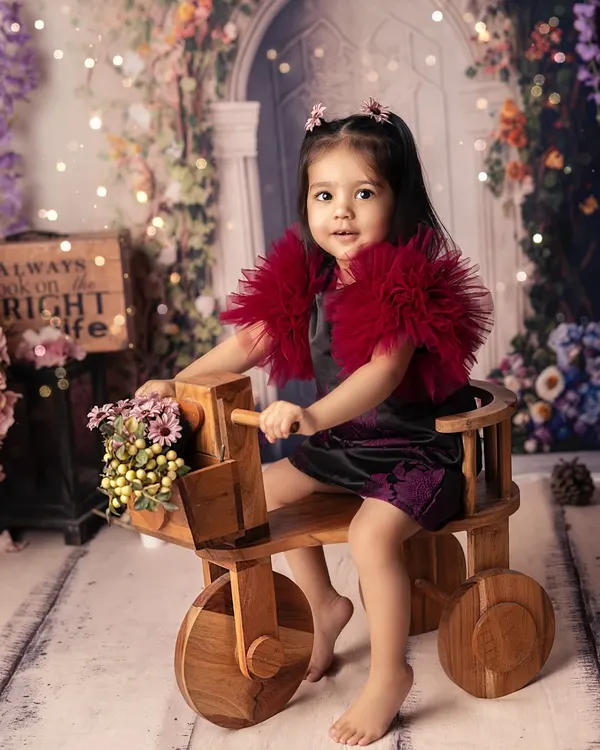 Pedaling through a garden of dreams on her little wooden trike. This whimsical setup, with its floral backdrop and soft lighting, creates a magical, fairytale-like portrait.