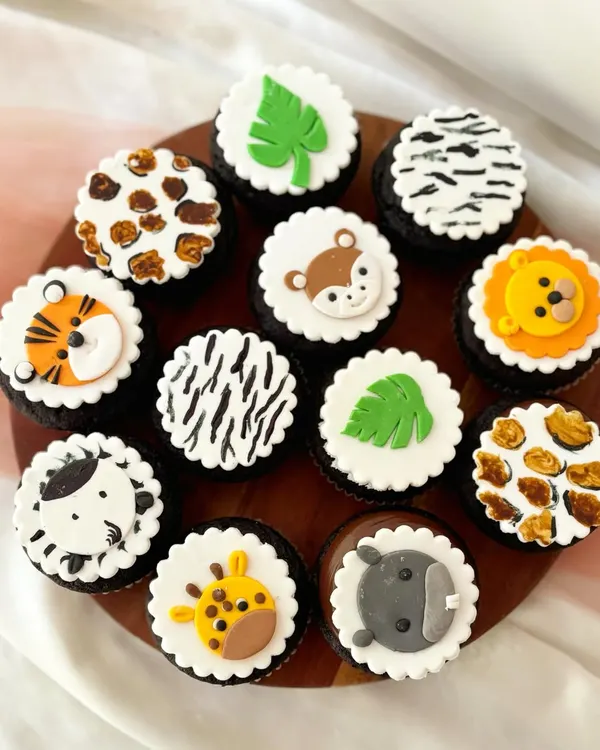 A dozen jungle safari cupcakes on rich chocolate bases. This set features a mix of fondant animal faces like a tiger, giraffe, and zebra, alongside animal print designs and monstera leaves for a complete jungle party theme.