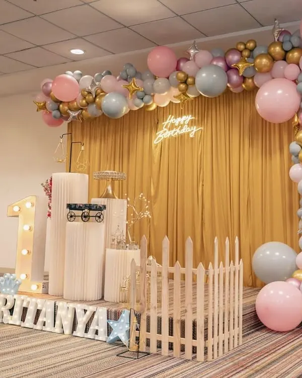A wider view of the elegant first birthday setup for Ritanya. The combination of a golden curtain backdrop, a neon sign, and a full balloon arch creates a rich and celebratory feel for the main photo area.