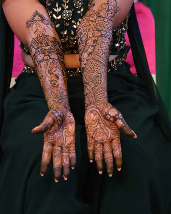 A beautiful example of a full bridal mehndi after the stain has set. The design covers both arms with a rich, dark color, featuring a mix of portraits, traditional patterns, and personalized story elements.