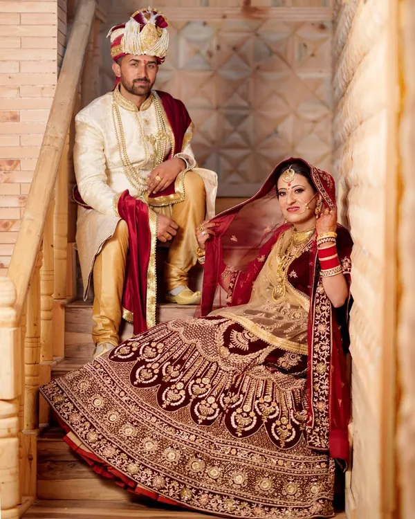 An elegant, royal-style wedding portrait of the couple posed on a wooden staircase. The bride's rich, velvet lehenga and the groom's sherwani create a timeless and regal look.