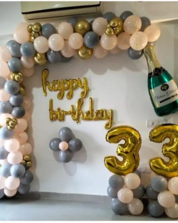 A classy 33rd birthday decoration with a balloon arch in grey, peach, and gold. The setup includes "Happy Birthday" foil letters and a champagne bottle balloon.