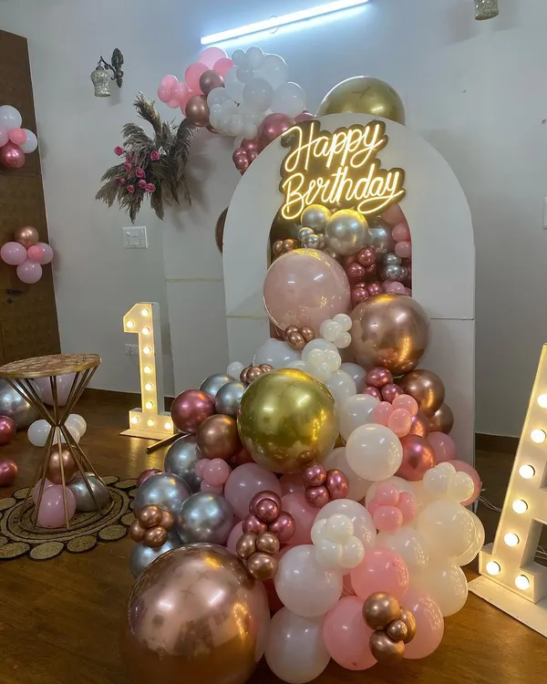 A beautiful and modern first birthday setup with a boho-chic feel. This decoration uses a cascade of rose gold, gold, silver, and white balloons flowing from an arched backdrop, complemented by a neon sign and marquee initials.