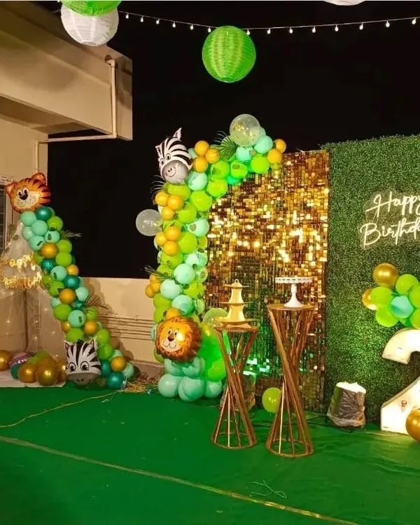 An exciting outdoor jungle safari theme for a second birthday. The setup includes a gold shimmer wall, a faux grass backdrop with a neon sign, a light-up number "2", and a balloon garland with animal accents.