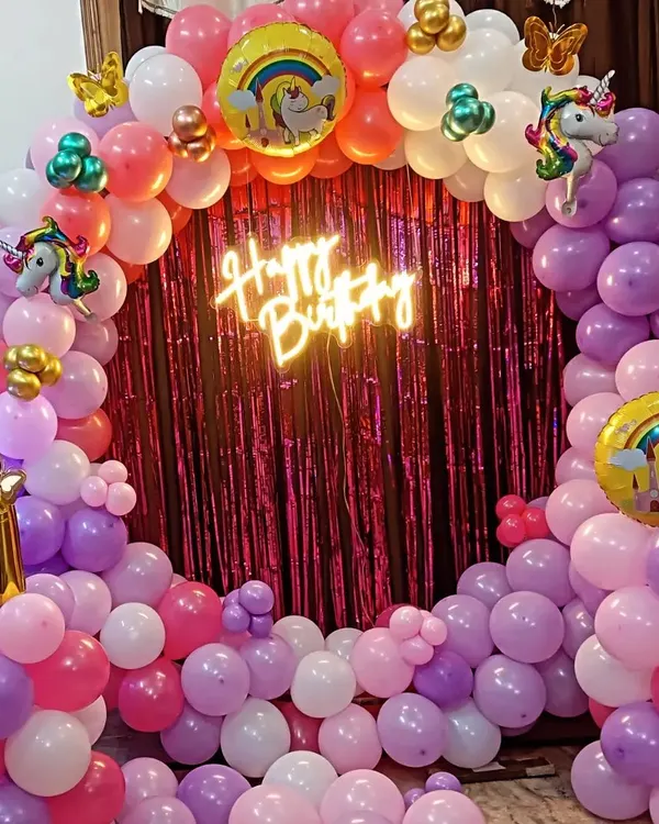 A magical Unicorn theme for a first birthday, featuring a circular balloon arch in pastel pinks and purples. The setup is completed with a "Happy Birthday" neon sign and unicorn-themed foil balloons.