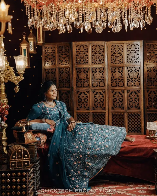A regal solo portrait of the bride-to-be in a stunning blue lehenga, seated on an ornate set with rich, warm lighting.