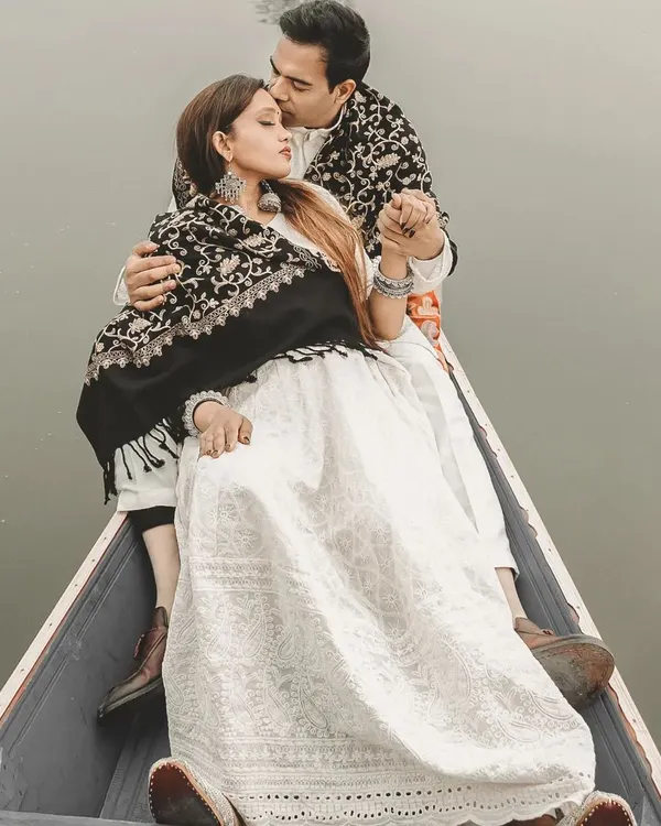 A close-up, tender moment on a Shikara boat in Kashmir. The groom-to-be wraps his partner in a traditional shawl, creating a warm and intimate portrait against the cool, misty backdrop of Dal Lake.