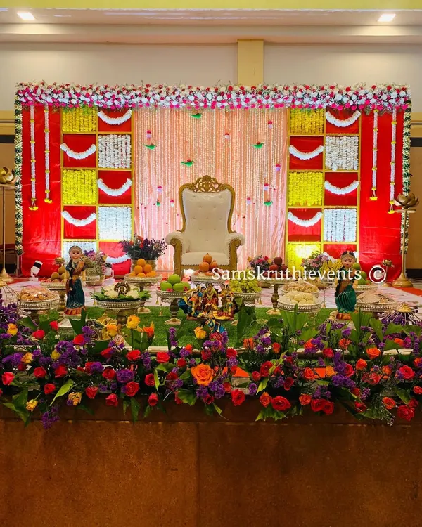 A traditional Seemantha decoration with a vibrant red and yellow checkered backdrop. The stage is beautifully framed with a thick border of fresh flowers.