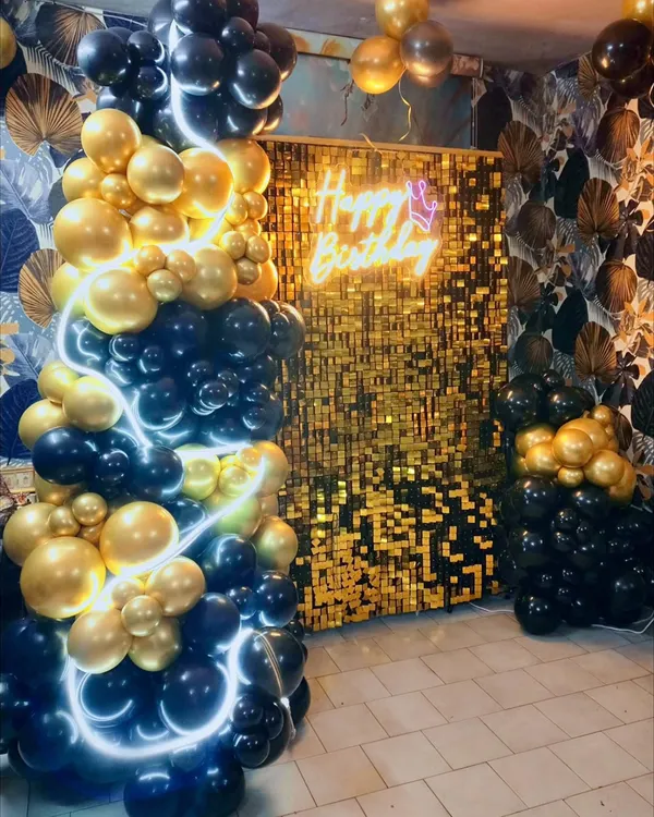 Glamour and sophistication for a memorable birthday. This stunning setup features a cascade of black and gold balloons, a glittering gold sequin backdrop, and a 'Happy Birthday' neon sign for a truly luxurious feel.