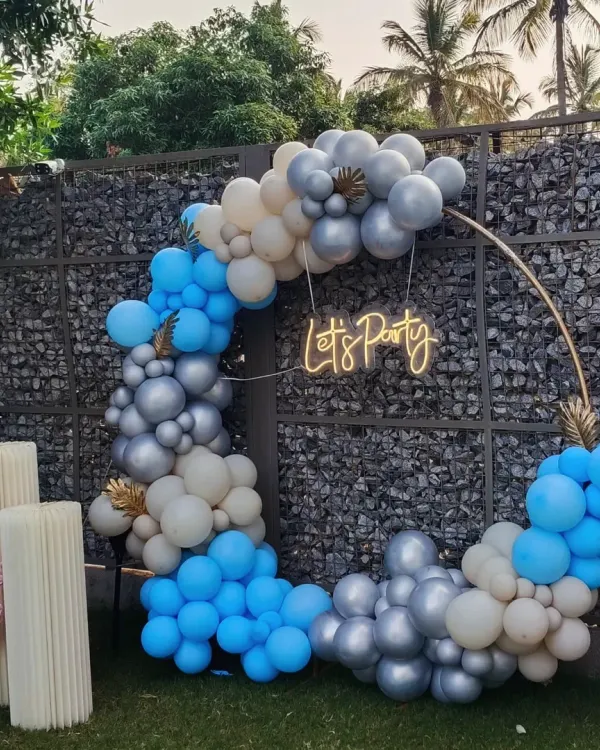 A chic and modern "Let's Party" setup perfect for any celebration. This outdoor decor features a circular frame with a blue, silver, and white balloon garland and a neon sign, creating a stylish photo spot.