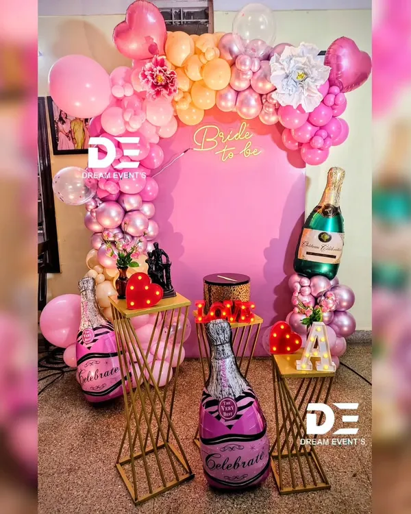 A fun and feminine "Bride to be" decor setup for a bridal shower. The pink backdrop is framed by a balloon arch, and the scene is accessorized with champagne bottle balloons and modern gold stands.