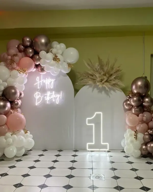 A stunningly modern first birthday setup in rose gold, blush, and white. The clean arched backdrops, neon sign, and organic balloon garlands create a sophisticated and trendy look.