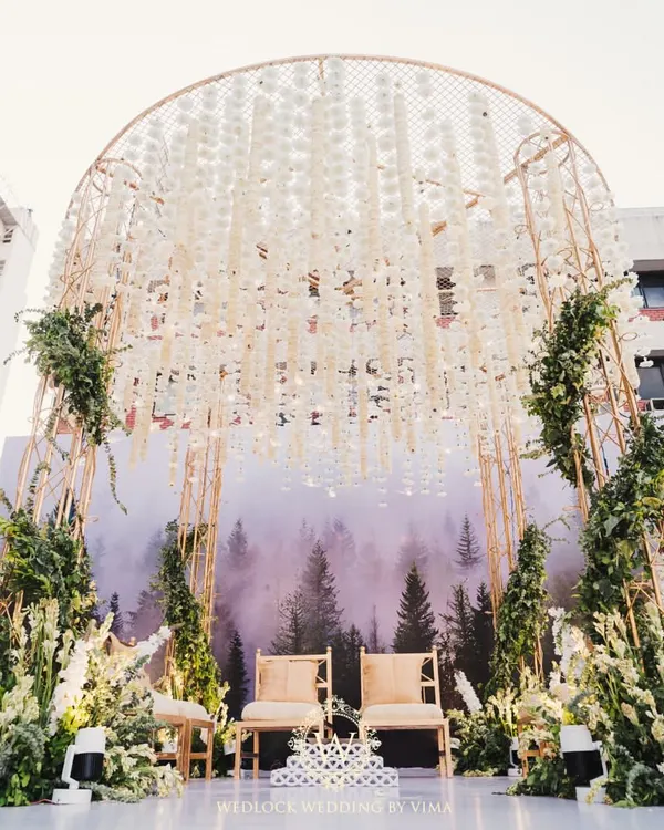 A modern minimalist mandap that perfectly blends nature and contemporary design. The structure features a backdrop of a misty forest, a canopy of hanging white florals, and lush green arrangements.