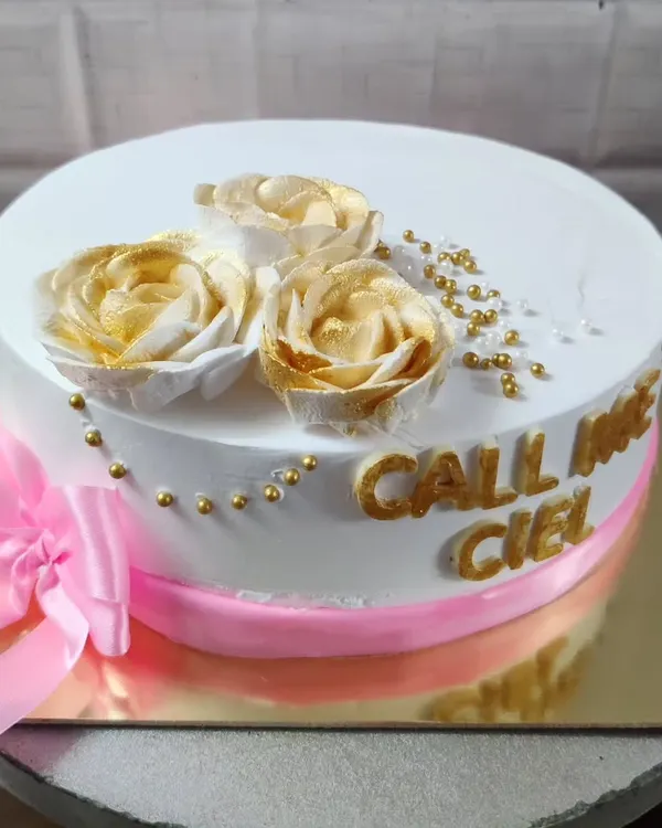 An elegant white cake with "Call me Ciel" written in gold. The design is finished with a pink ribbon and beautiful golden-dusted buttercream roses.
