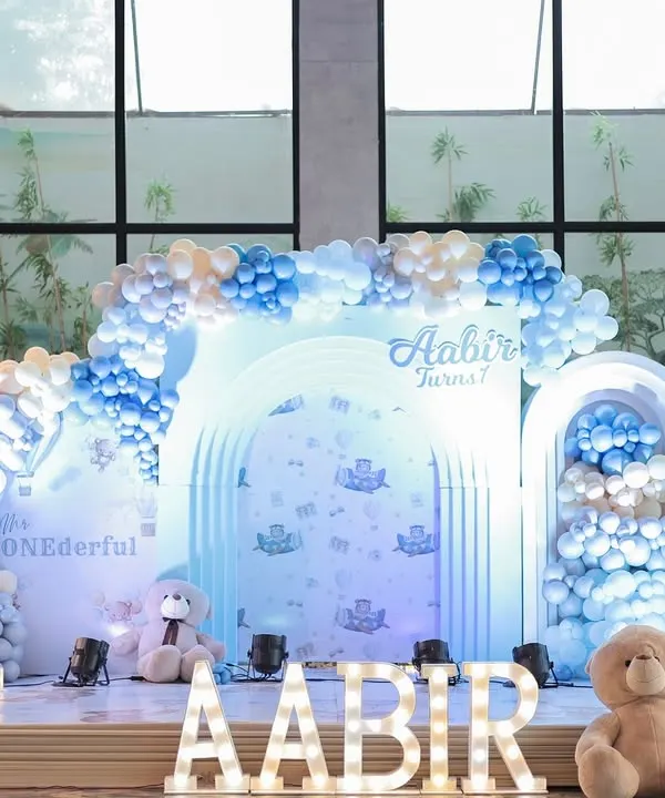 A full view of a dreamy blue and white teddy bear first birthday stage. The layered arches, cascading balloons, and large marquee letters create a stunning focal point for photos and celebrations.