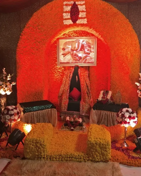 A vibrant Ganesh Chaturthi decoration. The gaddi is set inside a large arch made entirely of fresh orange and yellow marigolds, with a Tripundra symbol at the top, creating a bright and auspicious space for Ganpati Bappa.