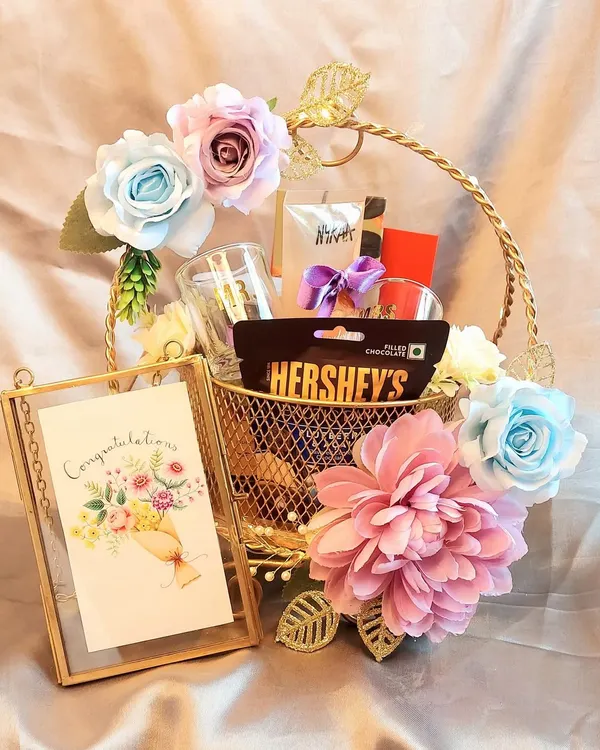 A sweet 'Congratulations' hamper to celebrate a special achievement. This golden basket is filled with chocolates, a personalized card in a glass frame, and beautiful pastel flowers.