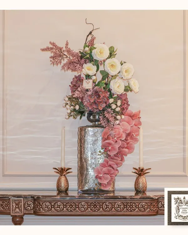 This console is styled with blooming elegance, featuring a dramatic floral arrangement in a tall silver vase. The display is flanked by two pineapple-shaped candle holders, adding a touch of tropical, traditional glow.