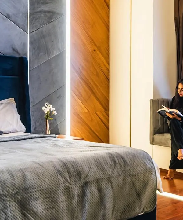 This master bedroom is designed as a cozy retreat. A comfortable reading nook is built into the bay window, and the deep blue velvet headboard is flanked by a geometric-paneled wall with brass inlays. The rich wood flooring and layered lighting create a warm, enveloping ambiance.