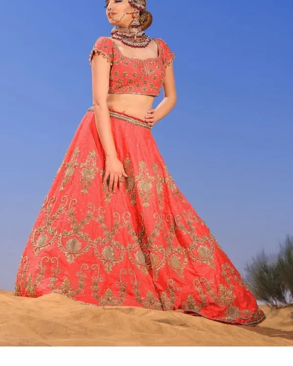A magnificent coral-red bridal lehenga, heavily embellished with intricate zardosi work. This is a statement piece for the bride who desires a grand and opulent look for her wedding day.