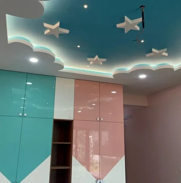 A playful and practical kids' bedroom featuring a custom wardrobe in pastel pink and teal. The highlight is the cloud-and-star-themed false ceiling with integrated lighting, creating a magical sky effect.