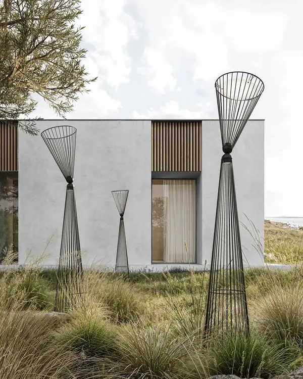 A grouping of Ikonika floor lamps by Karman in a landscaped garden. Their metal cage structure creates an airy visual volume that interacts beautifully with the surrounding grasses and modern architecture.